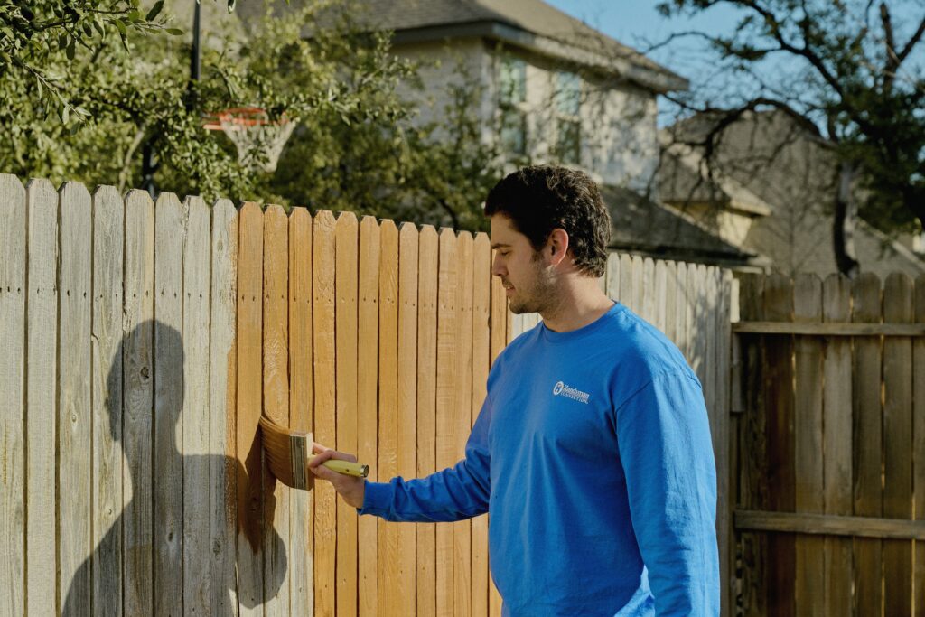 handyman using a paint brush to apply a stain to a wooden fence